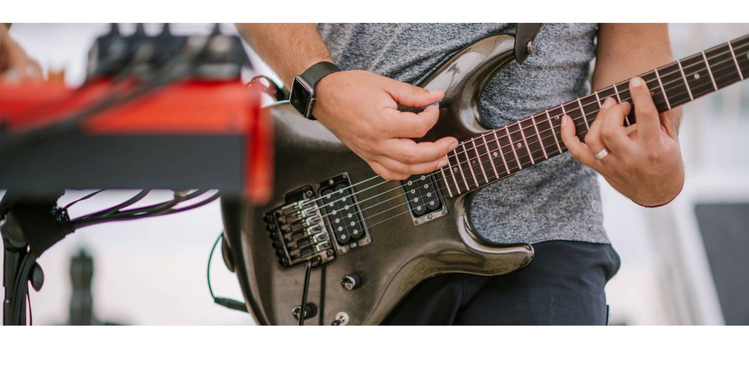 A close-up of a person in a grey shirt wearing a smartwatch, strumming a metallic charcoal grey electric guitar with an effects pedal board blurred in the foreground